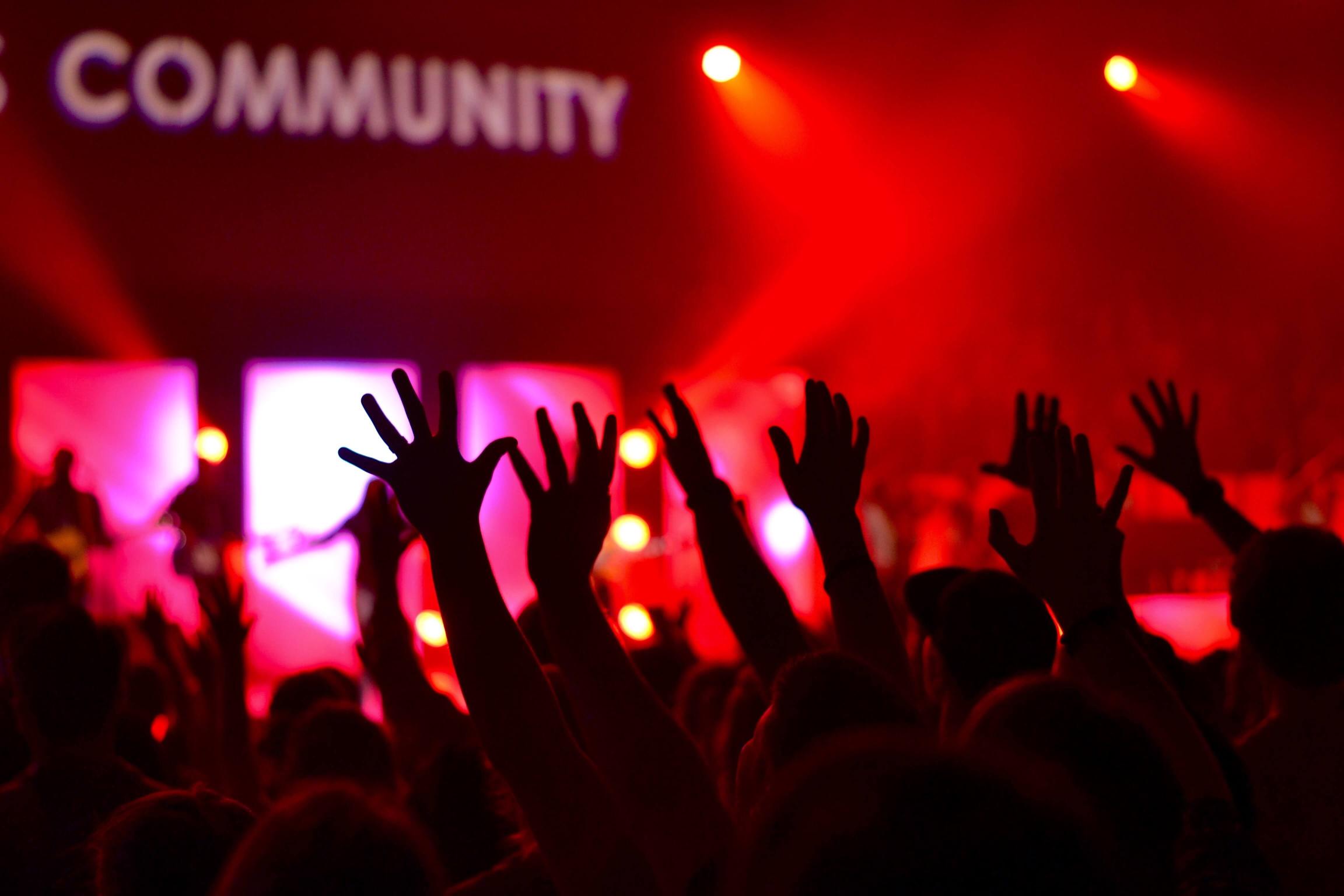 Man sieht die Hände einer feiernden Menschenmenge im rotem Licht. Das Wort 'Community' wird verschwommen an die Wand darüber projiziert. (c) Photo by William White on Unsplash Man sieht die Hände einer feiernden Menschenmenge im rotem Licht. Das Wort 'Community' wird verschwommen an die Wand darüber projiziert.